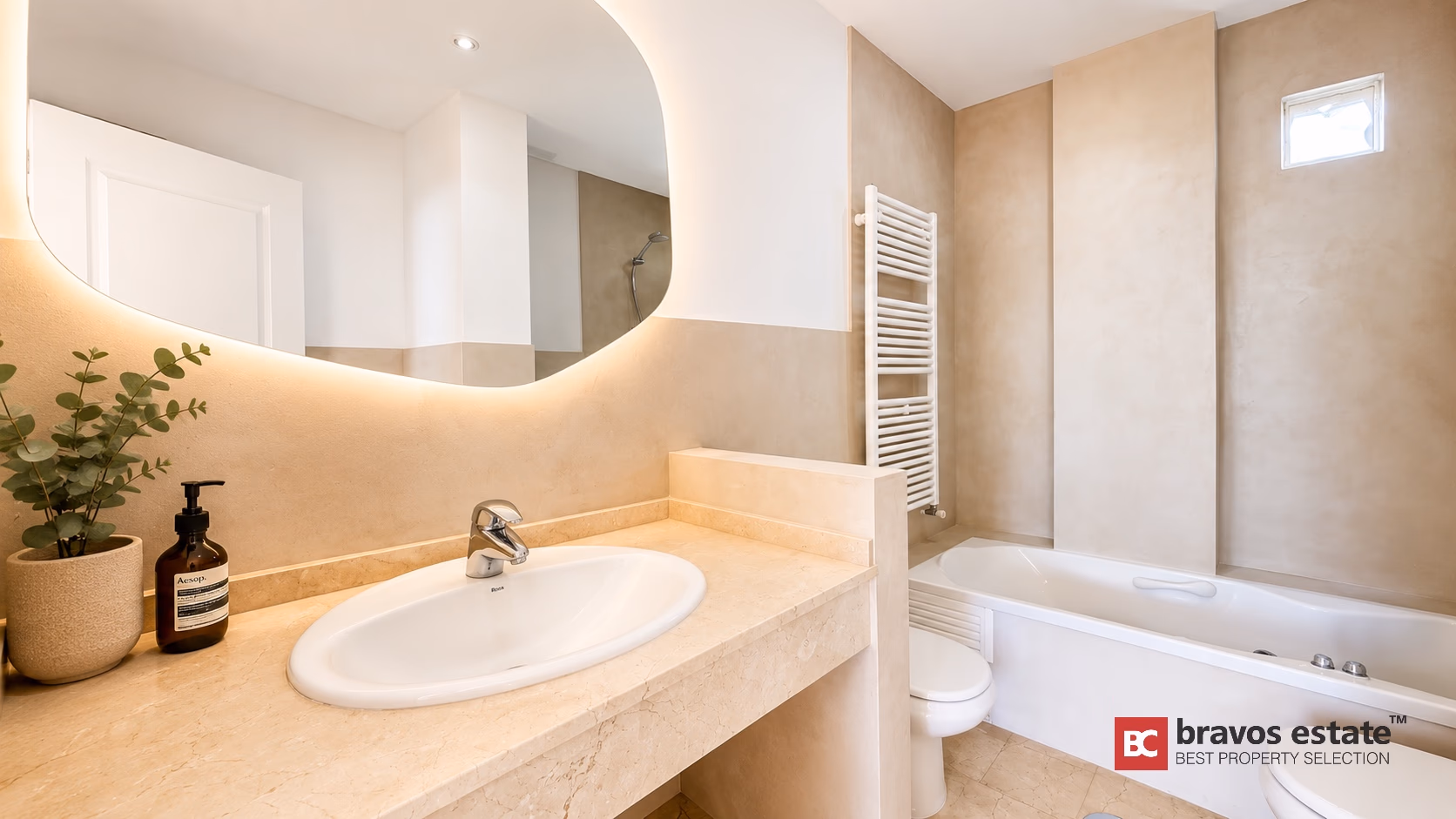 Salle de bain avec baignoire, miroir rétroéclairé, meuble vasque en pierre beige et murs carrelés