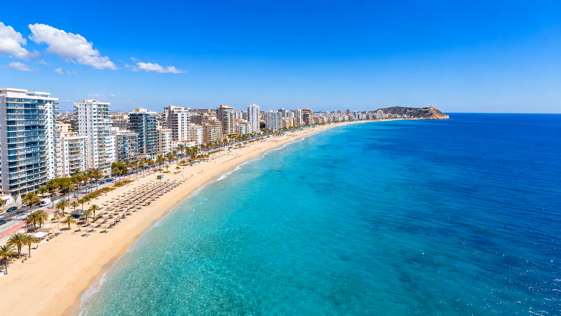 Luchtfoto van een strand aan de Costa Blanca in de provincie Alicante, met witte appartementengebouwen langs de boulevard en turquoise zeewater