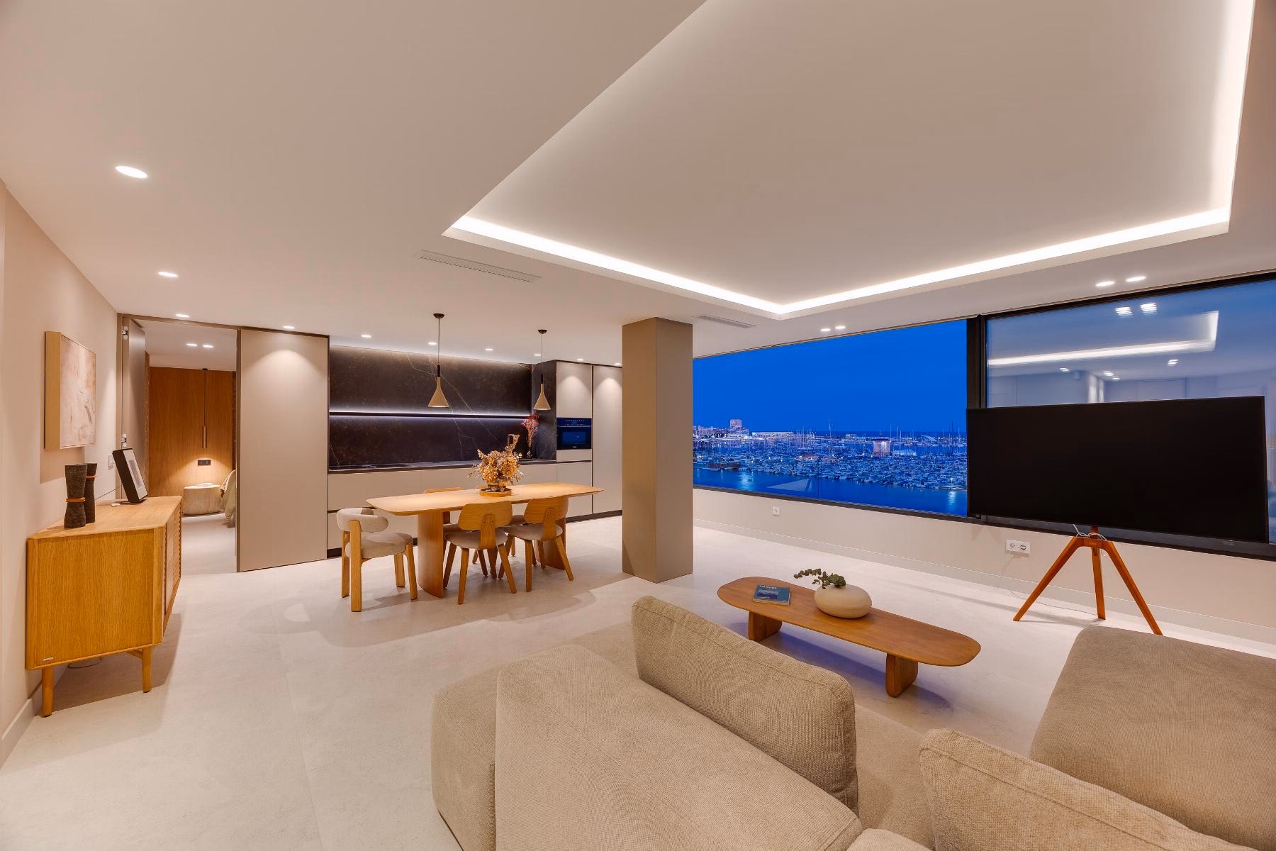 Open-plan living room with sofa, dining table and a wide window onto the sea at night