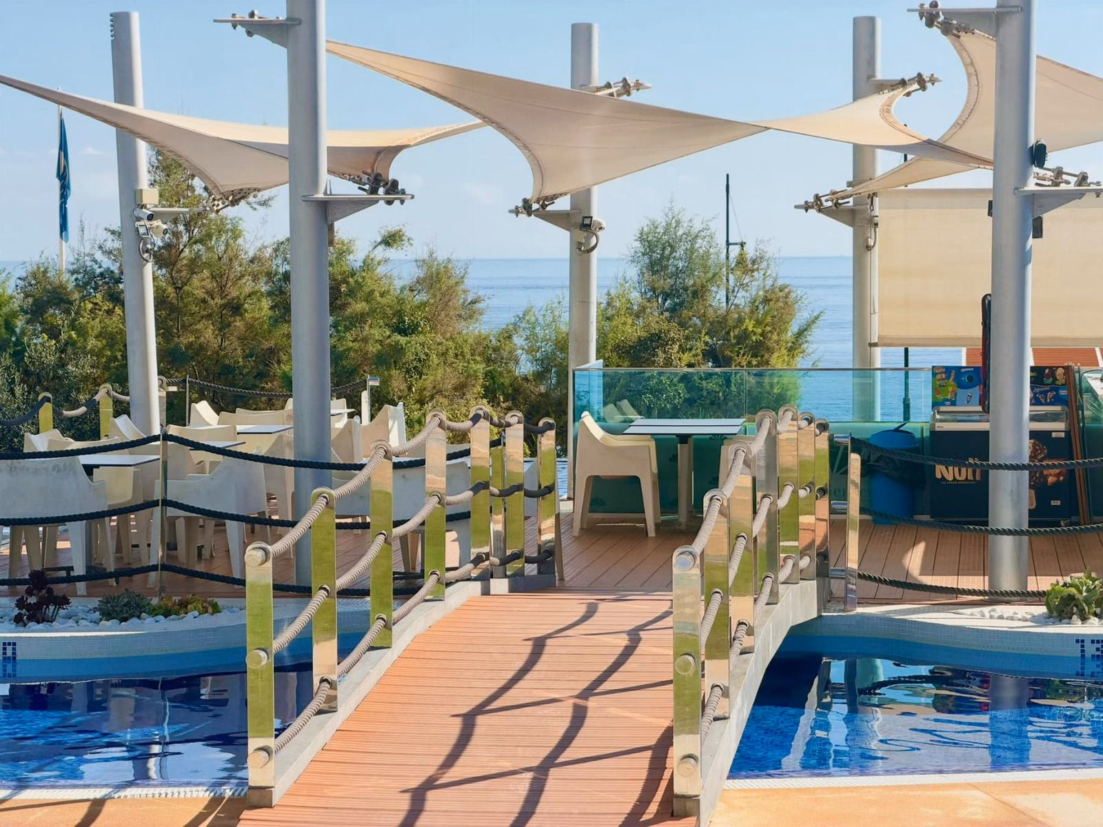 Piscina comunitaria con pasarela de madera y toldos blancos de vela en los jardines de Sea Senses