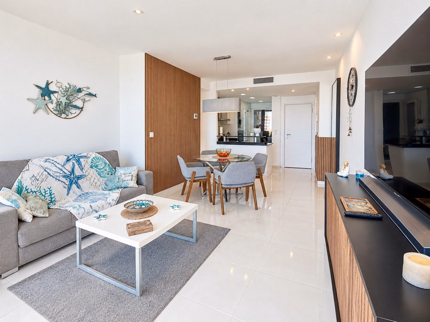 Salón con sofá gris, mesa de comedor y mueble de TV de madera sobre suelo blanco brillante