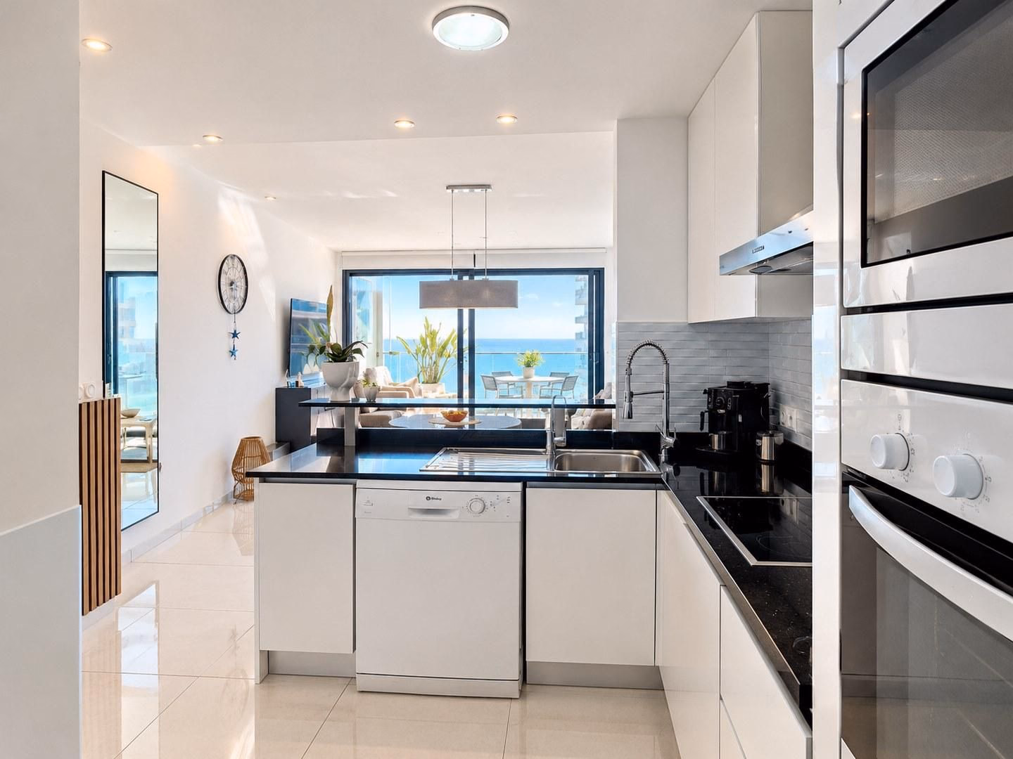 Cocina con muebles blancos y vista a través del salón hacia la terraza y el mar