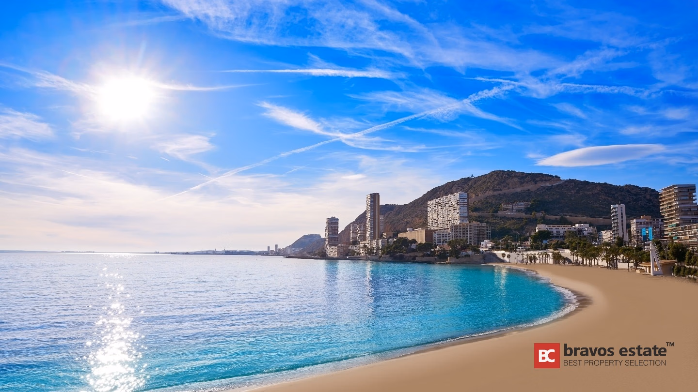 Playa de arena y mar Mediterráneo turquesa cerca de los apartamentos Bonalba Green, con el perfil costero de Alicante y una montaña rocosa bajo un cielo azul soleado.